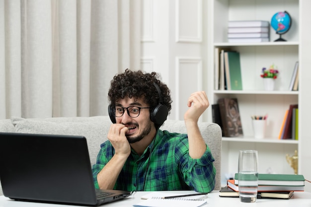 Studente online carino giovane ragazzo che studia sul computer con gli occhiali in camicia verde che morde il dito