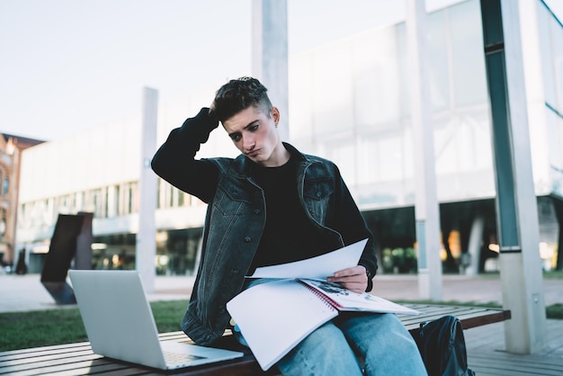 Studente premuroso con le carte in ginocchio guardando il computer portatile