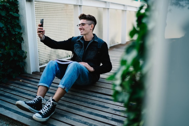 Studente sorridente con gli occhiali che si fa selfie mentre è seduto sul pavimento