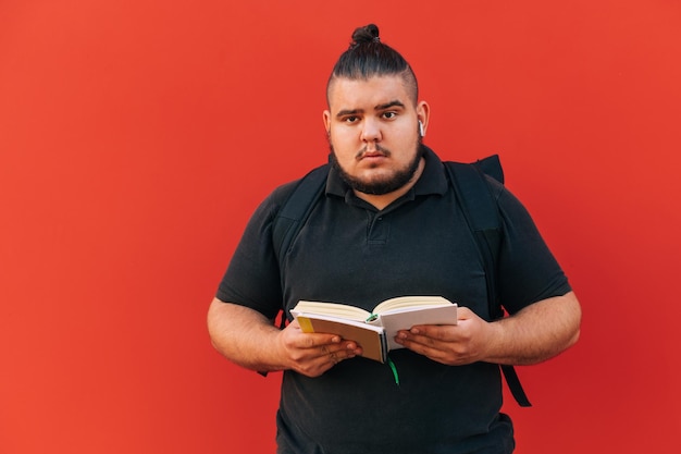 Studente sovrappeso divertente con uno zaino sulla schiena sta con un libro in mano su un muro rosso