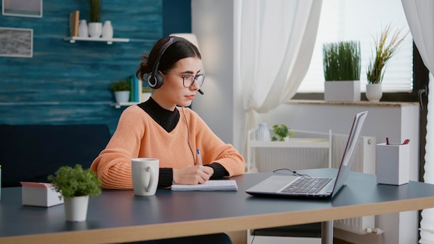 Studente universitario che ascolta la classe scolastica online sul laptop, utilizzando le cuffie in teleconferenza remota. Giovane che lavora su un corso web e prende appunti per studiare per conoscenza.