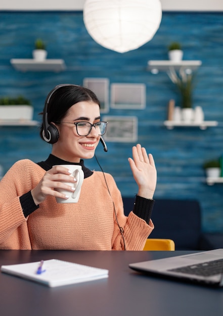 Studente universitario con bicchieri e tazza di caffè in mano saluta il suo insegnante durante la riunione in videochiamata parlando di laurea. Adolescente con le cuffie in testa seduto al tavolo della scrivania