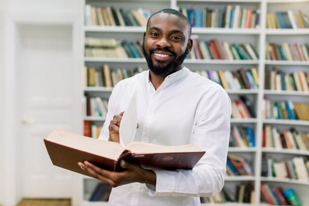 Studente universitario maschio afroamericano allegro sorridente in piedi nella moderna sala di lettura della biblioteca del college, tenendo il libro aperto