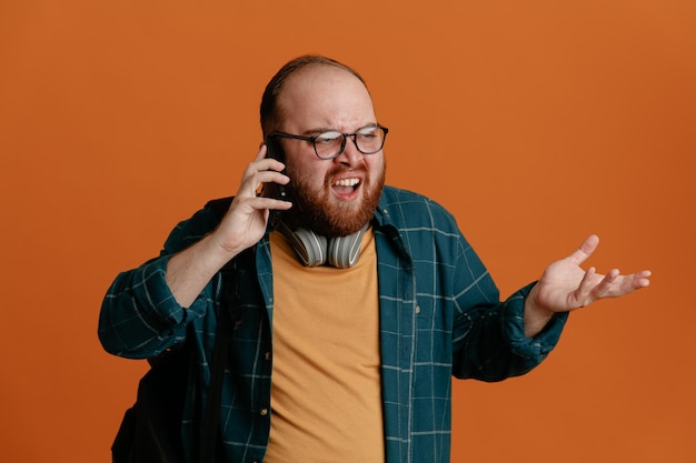 Studente uomo in abiti casual con zaino con le cuffie che parla al cellulare essendo arrabbiato e frustrato in piedi su sfondo arancione