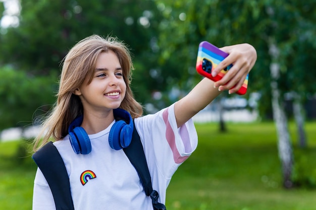 Studentessa adolescente caucasica che usa smartphone con custodia arcobaleno che si fa selfie all'aperto nel parco cittadino