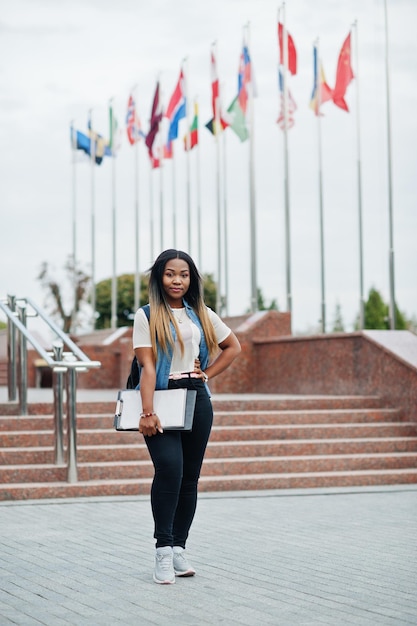 Studentessa africana in posa con zaino e articoli per la scuola nel cortile dell'università contro le bandiere di diversi paesi