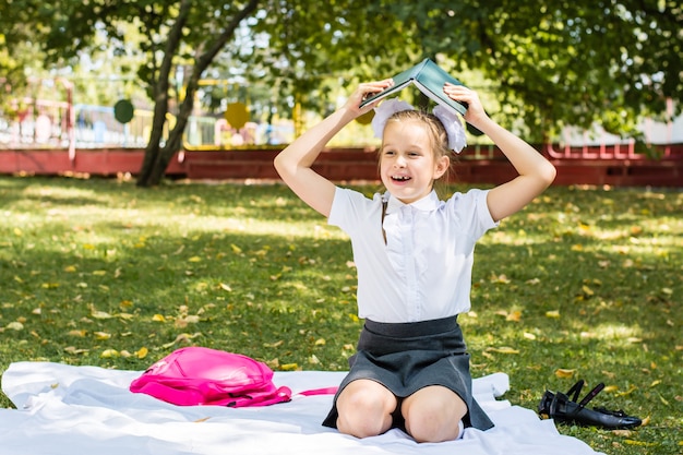 Studentessa allegra tiene un libro con i compiti sopra la testa mentre è seduta su una coperta in un soleggiato parco autunnale. Educazione all'aria aperta per i bambini. Ritorno al concetto di scuola