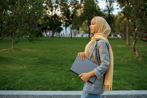 Studentessa araba con il computer portatile che cammina nel parco estivo. Donna musulmana che riposa sul sentiero a piedi.