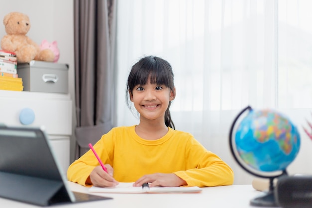 Studentessa asiatica che fa i compiti con il tablet digitale a casa I bambini usano i gadget per studiare Istruzione e apprendimento a distanza per i bambini Istruzione domiciliare durante la quarantena Resta a casa