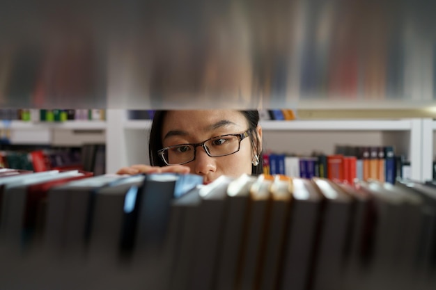 Studentessa asiatica che sceglie il libro di testo mentre si trova tra gli scaffali per libri nella biblioteca dell'università