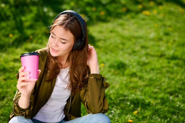 Studentessa attraente carina (donna) seduta rilassata sull'erba nel parco pubblico all'aperto e ascoltare musica in cuffia