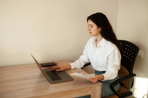 Studentessa attraente che studia la digitazione del testo utilizzando il laptop a casa Istruzione online e apprendimento a distanza Ragazza con i capelli ricci indossava una camicia bianca ufficiale studia online