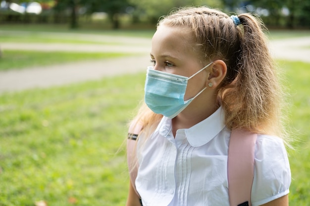 Studentessa bionda riccia in maschera protettiva in uniforme scolastica con zaino rosa torna a scuola