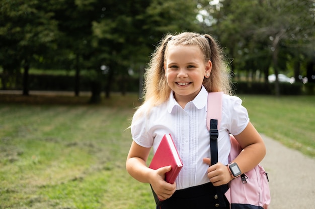 Studentessa bionda sorridente in uniforme scolastica che tiene il taccuino con zaino rosa torna a scuola supera