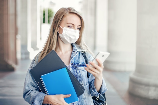 Studentessa che indossa una maschera antivirale protettiva legge i messaggi su un telefono cellulare durante la quarantena della pandemia di coronavirus