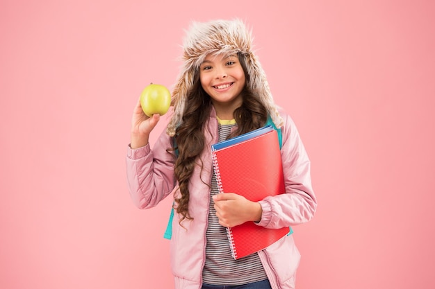 Studentessa con libri dopo la lezione pausa scolastica piccola ragazza felice paraorecchie cappello mangiare mela vacanze invernali vacanza adolescente bambino rosa sfondo allievo vita quotidiana educazione moderna studente del mese