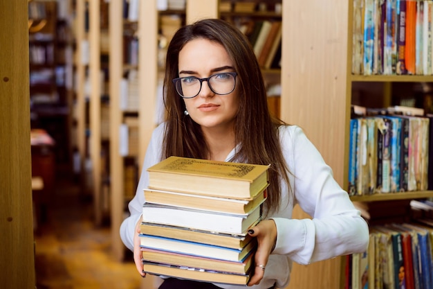 Studentessa con una grande pila di libri in biblioteca