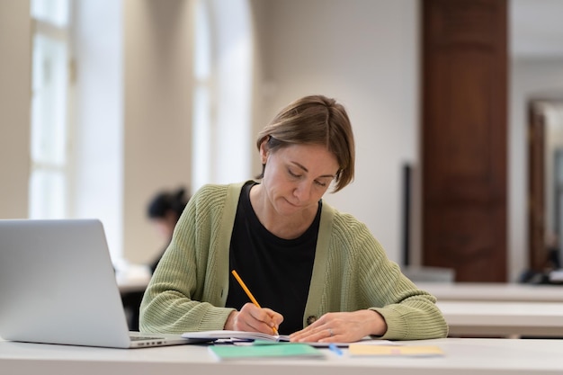 Studentessa di mezza età che si prepara per l'esame nella biblioteca pubblica mentre è seduta al tavolo con il laptop