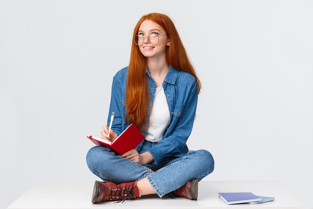 Studentessa diligente carina sognante e creativa con i capelli rossi, in bicchieri, seduto sul pavimento con le gambe incrociate circondato da quaderni scrivendo qualcosa