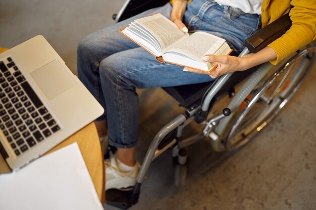 Studentessa disabile in sedia a rotelle tiene un libro, vista dall'alto, disabilità, interno della biblioteca universitaria sullo sfondo. Donna disabile che studia al college, le persone paralizzate acquisiscono conoscenza