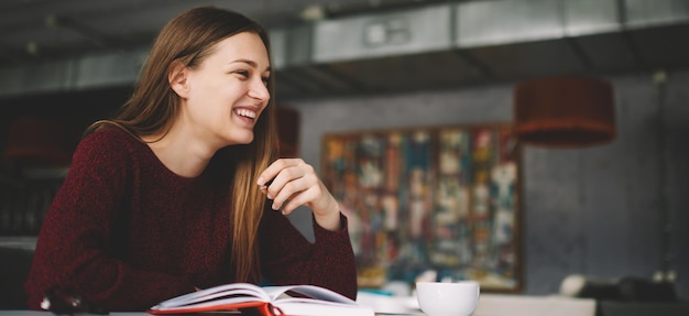 Studentessa felice e qualificata attraente che si siede al caffè accogliente che sorride spensierato alla rottura