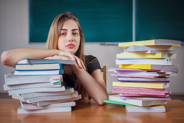 Studentessa frustrata che si siede allo scrittorio con un mucchio enorme dei libri di studio in aula.