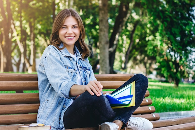 Studentessa giovane seduto su una panchina nel parco con libri, quaderni e cartelle. La ragazza insegna lezioni nel parco.