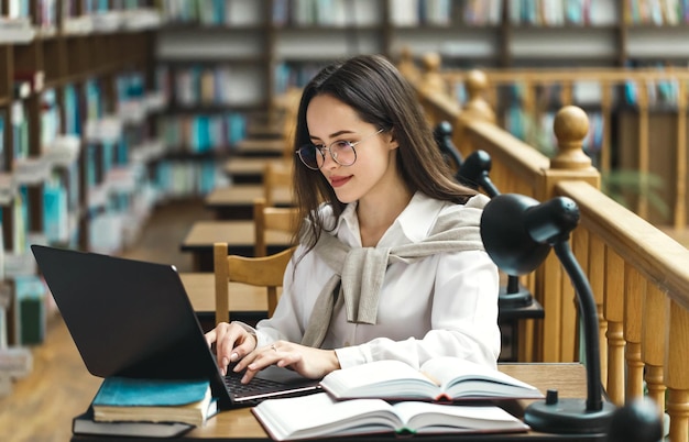 Studentessa graziosa con laptop e libri che lavorano in una biblioteca del liceo