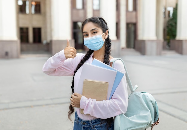 Studentessa indiana che indossa una maschera facciale e fa gesti con i pollici in su approvando il college in posa contro