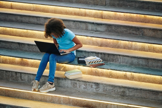 Studentessa intelligente che si siede sui gradini con molti libri in giro e lavora al computer portatile