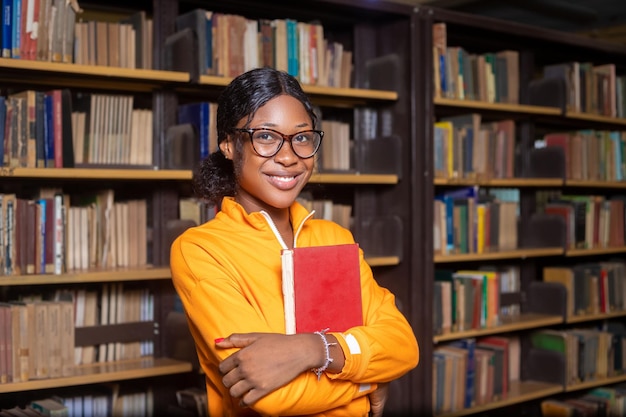 Studentessa internazionale allegra che sta accanto allo scaffale per libri nella biblioteca
