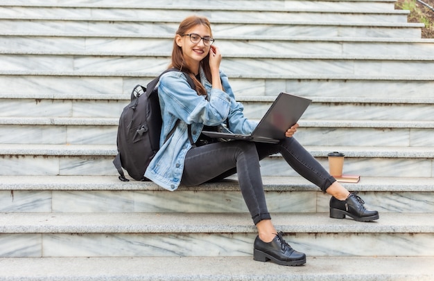 Studentessa moderna felice in rivestimento e zaino del denim che si siedono sulle scale con il computer portatile all'aperto. Insegnamento a distanza. Concetto di gioventù moderna.