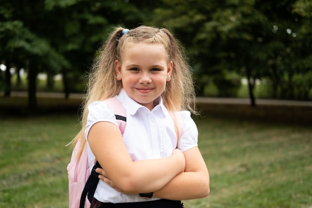 Studentessa riccia bionda felice in uniforme scolastica con zaino rosa torna a scuola all'aperto.