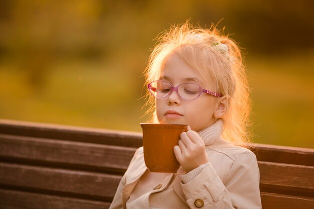 Studentessa riccia nei bicchieri e cappotto leggero con una grande tazza di cioccolata calda nel parco in autunno