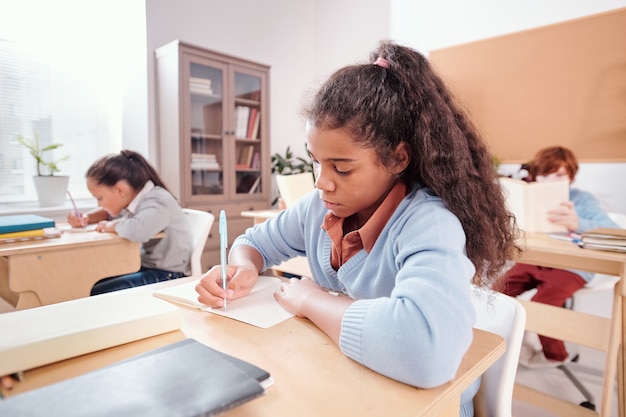 Studentessa seria di razza mista che prende appunti o scrive un saggio in un quaderno mentre è seduta alla scrivania a lezione e lavora individualmente