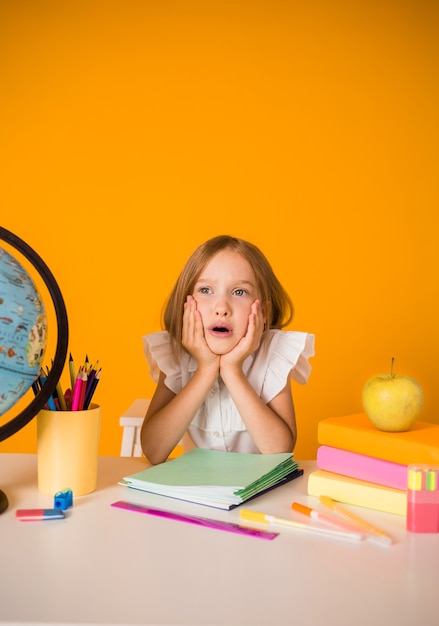 Studentessa sorpresa in una camicetta bianca è seduta a un tavolo con materiale scolastico e un globo e su uno sfondo giallo con una copia dello spazio. Orientamento verticale