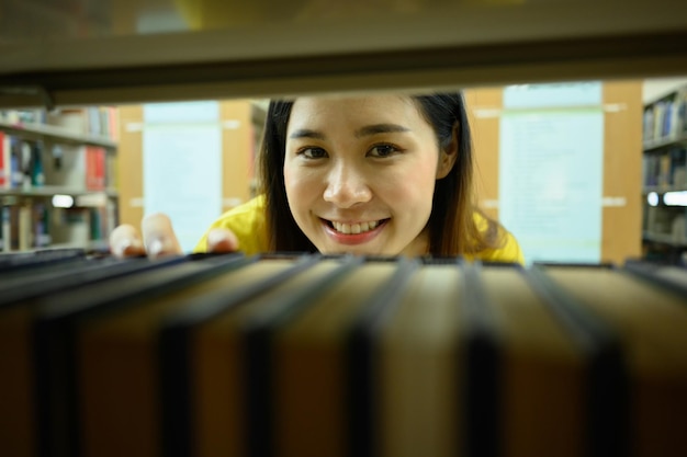 Studentessa universitaria asiatica allegra che sorride guardando attraverso uno scaffale in una biblioteca Apprendimento dell'istruzione e concetto di persone