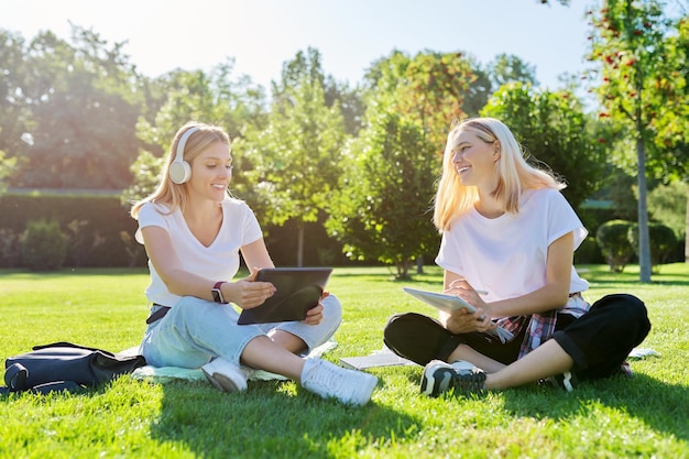 Studenti adolescenti di ragazze che si siedono sul prato verde nel parco con la compressa digitale dello zaino