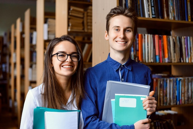 Studenti sorridenti maschi e femmine in biblioteca in piedi vicino a scaffali con libri, studi ed enciclopedie Agli studenti felici piace studiare