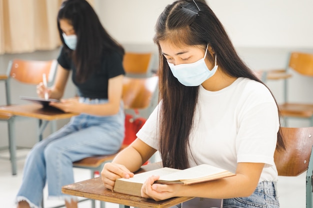 Studenti universitari asiatici tornano a scuola con la maschera facciale e mantengono la distanza sociale mentre studiano in classe per prevenire la pandemia di COVID-19