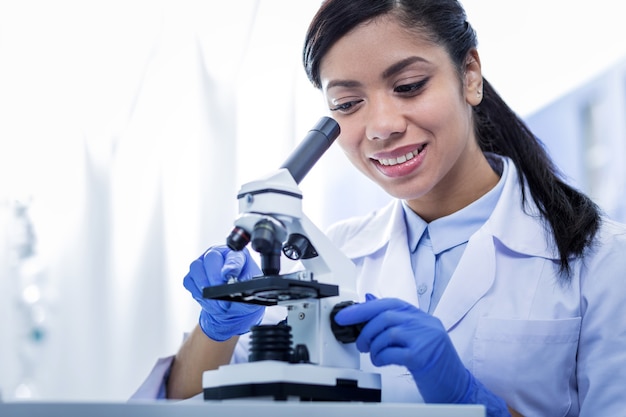Studi scientifici. Bella donna allegra positiva che indossa un camice ed esamina il microscopio mentre si lavora nel laboratorio scientifico