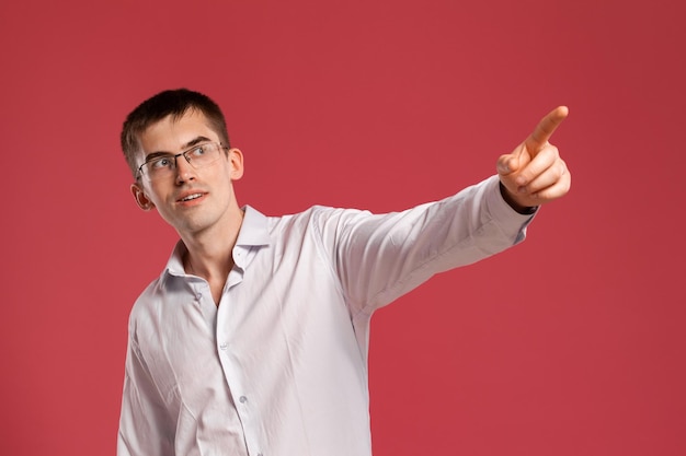 Studio di un giovane intelligente con una classica camicia bianca, orologi neri e occhiali che puntano a qualcosa mentre posano su uno sfondo rosa. Taglio di capelli alla moda. Concetto di emozioni sincere. Copia spazio.