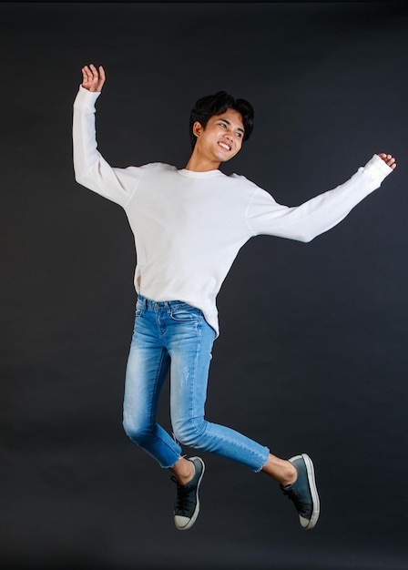Studio shot del giovane asiatico LGBTQ gay allegro felice bisessuale modello maschile omosessuale in abito casual saltando in alto in aria sorridente ridendo in posa tenendo due dita segno di pace su sfondo nero