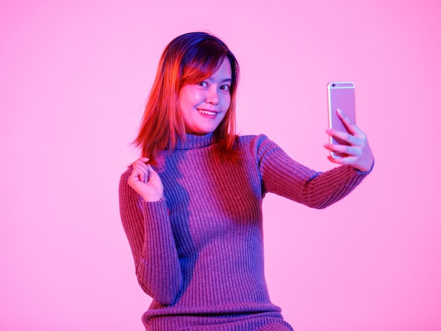 Studio shot del modello femminile alla moda urbano asiatico in maglione a collo alto grigio in piedi sorridente utilizzando smartphone su sfondo rosa chiaro.