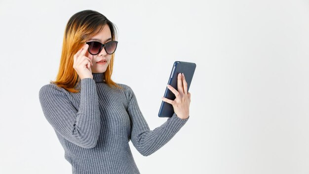 Studio shot del modello femminile asiatico giovane capelli tinti in maglione a collo alto grigio in piedi tenendo la mano toccando moda occhiali da sole lenti nere guardando il monitor tablet touchscreen su sfondo bianco.