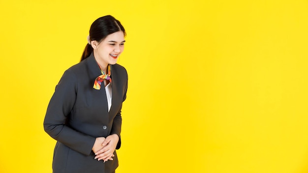 Studio shot di assistente di volo femminile di successo professionale asiatica hostess in uniforme formale che indossa bretelle denti in piedi sorridente inchinandosi a testa in giù saluto passeggero su sfondo giallo.