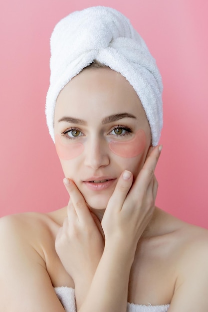 Studio shot di donna caucasica soddisfatta e lentigginosa che indossa un asciugamano bianco sulla testa con macchie di collagene sotto gli occhi in piedi nuda su sfondo rosa Concetto di prodotto cosmetico per la cura della pelle