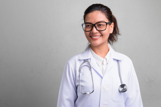 Studio shot di giovane bella donna asiatica medico indossando occhiali da vista contro il bianco