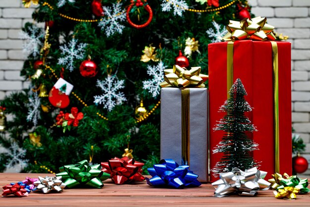 Studio shot di scatole regalo alte rosse e grigie con nastro dorato lucido posto su tavolo di legno con farfallino lucido colorato davanti bellissimo albero di pino di vigilia di Natale decorativo in sfondo sfocato.