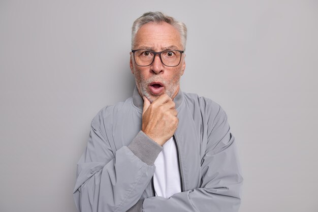 Studio shot di stordito barbuto dai capelli grigi uomo anziano tiene il mento senza parole